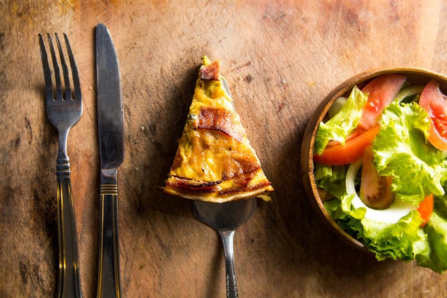 Part de quiche dorée posée sur une planche en bois, avec une salade verte, une fourchette et un couteau.