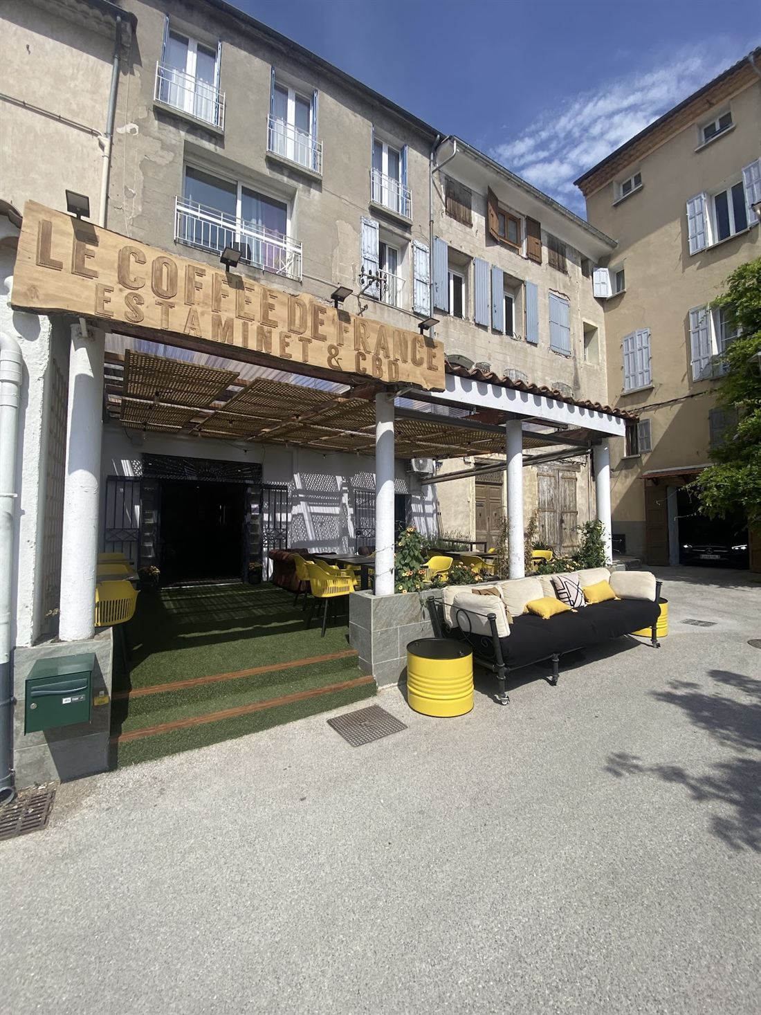 Façade du Coffee de France à Manosque : enseigne Estaminet, entrée sous auvent et terrasse avec tabourets jaunes.