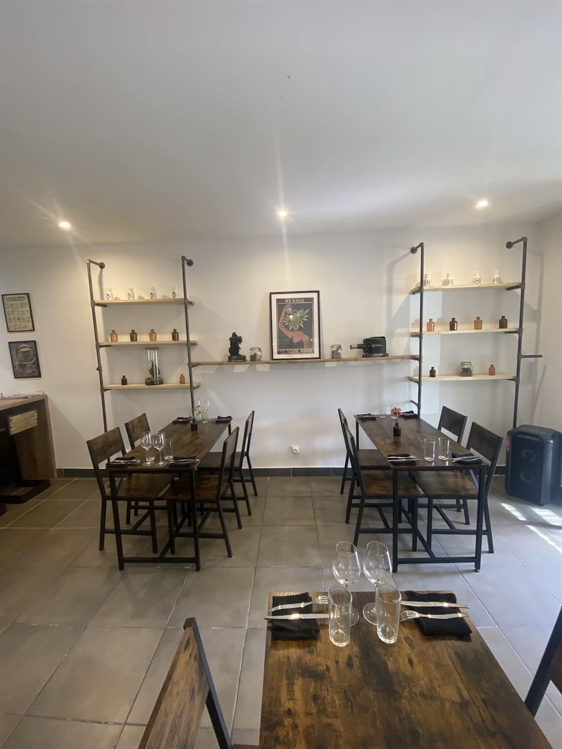 Salle du restaurant : tables en bois brut, chaises de bistrot noires et étagères industrielles.